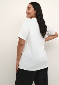 Kvinde med langt mørkt hår iført en ensfarvet hvid kortærmet T-shirt og sorte bukser, smilende og kigger til siden mod en lys baggrund.