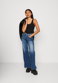 Top negro sin mangas, jeans de tiro alto y pierna ancha en azul, y zapatos negros de suela gruesa. La modelo posa con una mano en el bolsillo, con una chaqueta de cuero colgando del hombro.