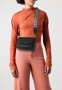Černá kožená crossbody taška s kulatým klopným víkem, texturovaným povrchem a dekorativním vzorovaným popruhem. Modelka má na sobě dlouhý oranžový top.