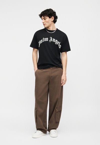 Νεαρός άνδρας κοιτάζοντας αριστερά, φορώντας μαύρο μπλουζάκι "Palm Angels", καφέ cargo παντελόνι, λευκά αθλητικά παπούτσια και ασημένιο κολιέ αλυσίδα.