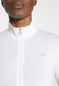 White zip-up jacket with a high collar, featuring a pink zipper and a small pink logo on the chest. Smooth fabric texture.