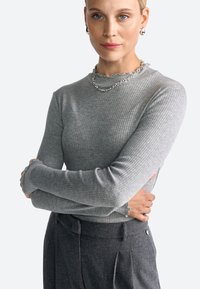 Szürke bordázott hosszú ujjú felső, fodros nyakkivágással és szűk fazonnal, sötét, fazonra szabott nadrággal párosítva, gombos részletekkel.