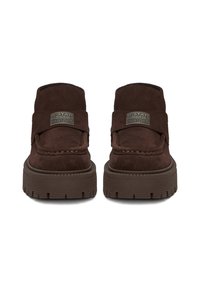 Zapatos de ante marrón tipo slip-on con suela gruesa de goma, que presentan detalles de costura y un parche de marca prominente en la parte superior.