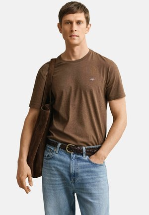 Ung mann iført en brun T-skjorte, lyseblå jeans og et flettet belte, som bærer en brun handlenettpose over skulderen.