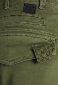 Pantalon en coton vert olive avec une finition texturée, comportant plusieurs poches cousues et une boucle tissée pour accessoires. Détail d'étiquette noire présent.