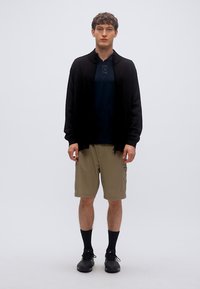 Μαύρο μακρυμάνικο ζακέτα πάνω από μπλε μπλούζα με στρογγυλή λαιμόκοψη, μπεζ σορτς καρό, μαύρες κάλτσες και μαύρα παπούτσια με κορδόνια.