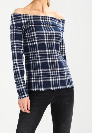 Žena v námořnicky modrém a bílém kostkovaném topu s odhalenými rameny a dlouhými rukávy, doplněném černými džínami, na jednobarevném pozadí.