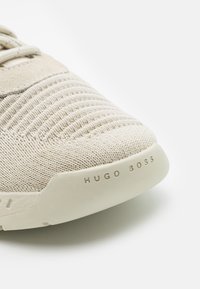 Beigen värinen neulottu tennari, jossa on teksturoitu päällinen, sileä pohja ja hillitty logotarroin sivussa. Kevyt ja minimalistinen muotoilu.