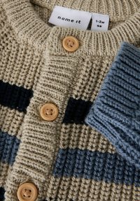 Beżowy sweterek dla niemowląt wykonany na szydełku, z niebieskimi i czarnymi paskami, z drewnianymi guzikami oraz widoczną etykietą rozmiaru "1-2M 56" wewnątrz kołnierza.