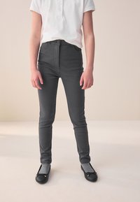 Pantalon gris taille haute, coupe ajustée, avec fermeture à boutons sur le devant et poches, associé à des ballerines noires et des chaussettes grises.