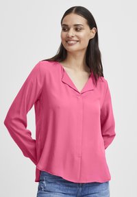 Pink long-sleeve blouse featuring a V-neckline and a curved hem. Made from a smooth fabric with a slight sheen and minimal detailing.