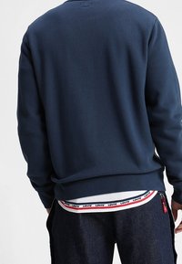 Námořnická modrá mikina s žebrovanou strukturou, kulatým výstřihem a dlouhými rukávy, doplněná bílým tričkem s červeným logem na spodním lemu.