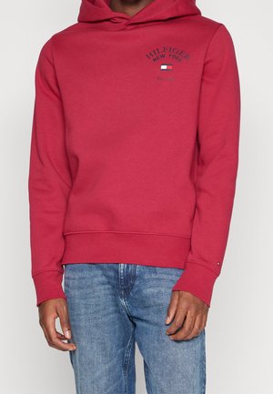 Osoba ubrana w czerwoną bluzę z kapturem Tommy Hilfiger z logo na piersi i niebieskie dżinsy, stojąca na jednolitym tle.