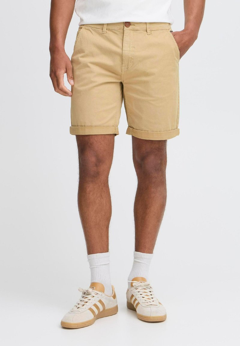 Pantalones cortos de algodón beige con dobladillos enrollados, que cuentan con un cierre de botón y cremallera frontal, llevados con calcetines blancos y zapatillas beige.