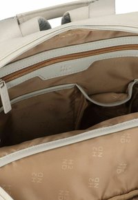 Interno di uno zaino in stoffa beige chiaro con foderatura beige, dotato di un compartimento principale, una tasca con zip e ulteriori dettagli di tasche più piccole.