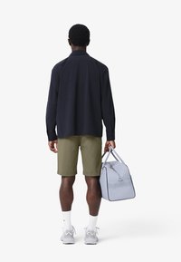 Chemise boutonnée bleu marine, shorts olive, baskets grises, chaussettes blanches ; tenant un sac gris clair avec des accents perforés.