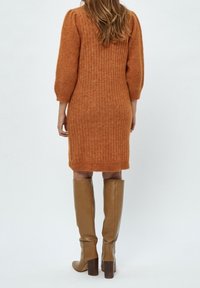 Femme portant une robe côtelée de couleur rouille, longueur genoux, avec des manches bouffantes et des bottes en cuir marron clair montant jusqu'aux genoux, vue de dos.