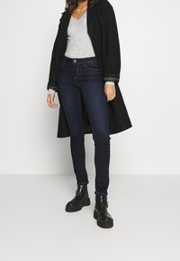 Černý vlněný kabát, šedý svetr s výstřihem do V, tmavé slim džíny a černé kotníkové boty. Oversized kabát s velkými knoflíky, přiléhavý design a hladká textura.