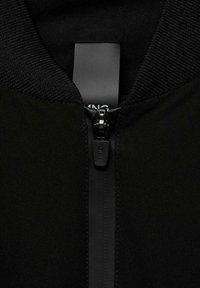 Musta vetoketjullinen takki, jossa on sileä, mattapintainen rakenne ja korkea kaulus. Vetoketjun vedin on varustettu hillityllä brändilogolla. Kangas näyttää pehmeältä ja joustavalta.