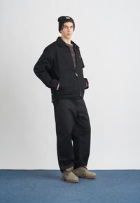 Μαύρο φερμουάρ μπουφάν με γιακά, επάνω από ένα ριγέ πουκάμισο. Μαύρες φαρδιές παντελόνες και ανοιχτό γκρι μποτάκια με χοντρές σόλες. Μαύρο σκουφάκι.