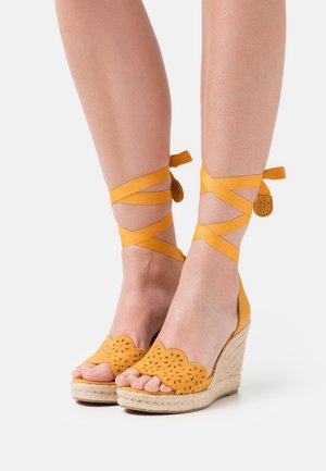 Sandales compensées jaunes avec un bord ondulé et un motif floral découpé, avec des rubans à la cheville et un talon enveloppé de jute.