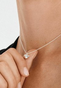 Sidabrinė vėrinys su širdies pakabuku, blizgios paviršiaus ir subtilios grandinėlės, demonstruojamas ant odos.