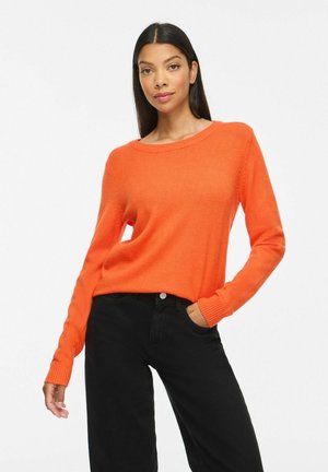 Femme aux longs cheveux foncés portant un pull orange et un pantalon noir, posant avec une main dans la poche devant un fond blanc.