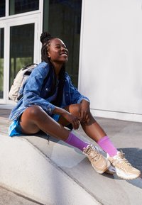 Giovane donna con giacca di jeans, gonna blu, calze viola e sneakers beige sorride seduta su un bordo di cemento all'aperto con uno zaino bianco.