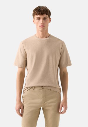 Mężczyzna w beżowej dzianinowej koszulce z krótkim rękawem i beżowych spodniach, stojący na tle jednolitego jasnoszarego tła.