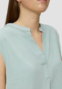 Lichtblauwe, mouwloze top gemaakt van een getextureerde stof. Beschikt over een kleine kraag en een knooploze placket met stikdetails.