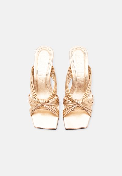 Par zlatnih sandala s tankim remenčićima, otvorenih na prstima s čvor detaljem i kvadratnim vrhom, prikazanih iz ptičje perspektive na jednostavnoj pozadini.