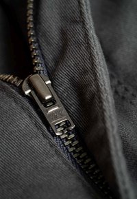 Schwarzer Stoff mit einer Nahaufnahme eines Metallreißverschlusses, der einen rechteckigen Schieber und Zähnchen-Details aufweist, vor einem strukturierten Hintergrund.