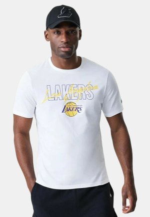 Uomo che indossa una maglietta bianca dei Los Angeles Lakers con logo giallo e viola e un cappellino nero dei Lakers, in piedi davanti a uno sfondo semplice.