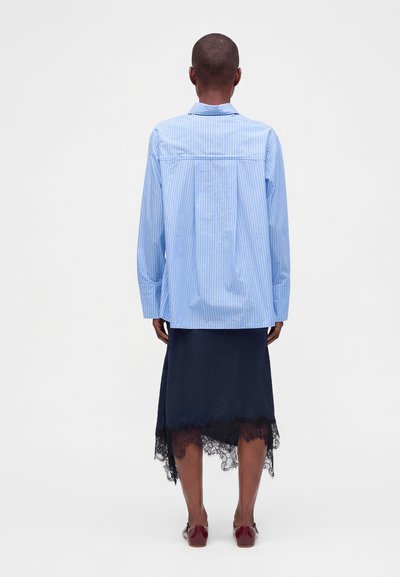 Personne portant une chemise à manches longues rayée bleue, une jupe bleu marine à ourlet en dentelle et des chaussures rouges, debout de dos devant un fond uni.