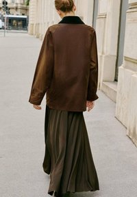 Person med hår i en knold, der går på en fortov i byen iført en brun jakke og en lang mørk nederdel ved beige bygningers vægge.