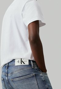 Camiseta blanca de algodón combinada con jeans de mezclilla azul. Los jeans cuentan con una etiqueta visible de Calvin Klein Jeans en la cinturilla.