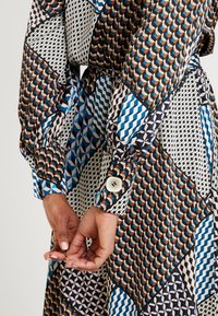 Langsleeve jurk met een patchworkontwerp met geometrische patronen in blauw, bruin en zwart, met knoopmanchetten en een zachte stofstructuur.