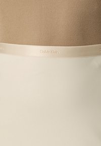 Gros plan sur un tissu beige côtelé au-dessus d'une ceinture lisse crème embossée avec le logo Calvin Klein dans une couleur assortie.