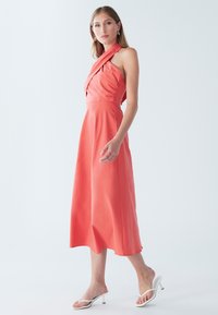 Korallpunane kaelakaelusega kleit mähitud peoosaga, voolava seeliku ja siledapinnalise tekstuuriga. Sobitatuna valgete rihmikandadega, esitatud neutraalsel taustal.