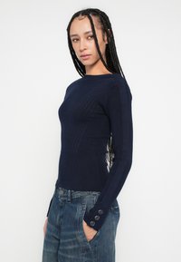 Námornícky modrý rebrovaný pletený sveter s dlhými rukávmi a gombíkmi na manžetách, priliehavý strih zvýrazňujúci trup, nosený s džínsami.