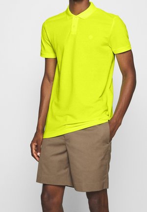Man draagt een felgeel poloshirt en beige korte broek, staat met één hand in de zak tegen een effen achtergrond.