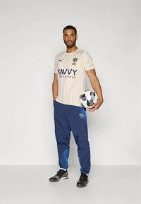 Puma sporta krekls ar bēšām svītrām un zilu logotipu, apvienots ar zilām biksēm ar zilām akcentiem. Tur rokās melnu un baltu futbola bumbu.