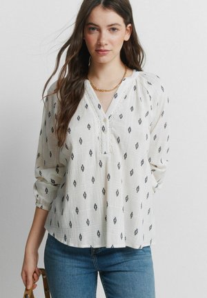 Nuori nainen, jolla on pitkät ruskeat hiukset, valkoinen kuvioitu paita ja siniset farkut, pitäen tan-värisen laukkuremmin yhdellä kädellä.