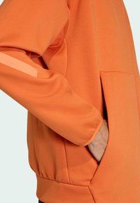 Felpa arancione con colletto alto, tasca laterale e una striscia riflettente a contrasto lungo la manica. Tessuto morbido e strutturato.