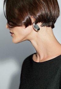Una donna con un hairstyle a caschetto corto indossa un grande orecchino moderno. Si volge leggermente di lato su uno sfondo neutro.