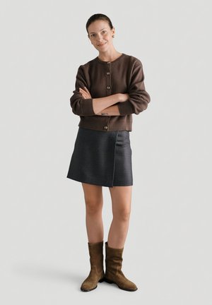 Kobieta o jasnej karnacji ubrana w brązowy kardigan na guziki, ciemną w kratkę spódnicę i brązowe zamszowe buty do połowy łydki, stoi z założonymi rękami.