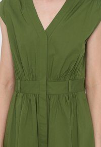 Femme portant une robe sans manches vert olive avec un col en V et une ceinture assortie à la taille.
