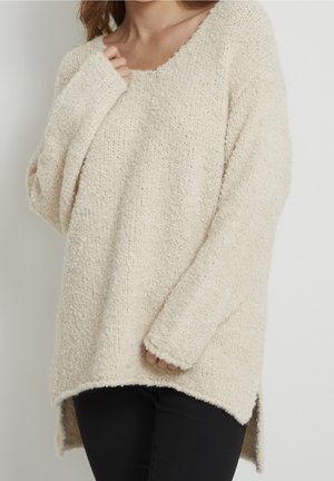 Osoba ubrana w kremowy, teksturowany sweter z długim rękawem, o dekolcie w szpic i asymetrycznym, dłuższym tyłem, połączony z czarnymi spodniami na jednolitym tle.