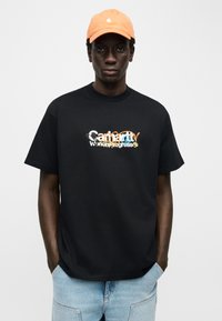 Νέος άνδρας που φοράει μαύρο t-shirt με σχέδιο και ανοιχτό μπλε τζιν, με τα χέρια στις τσέπες, πορτοκαλί καπέλο, σε απλό λευκό φόντο.