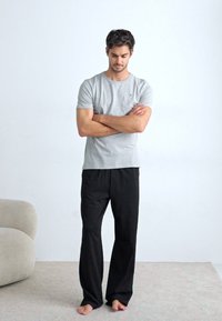 T-shirt grigia a maniche corte e pantaloni neri larghi; design semplice, materiale in cotone, indossati a piedi nudi, con le braccia incrociate in una posa neutra.
